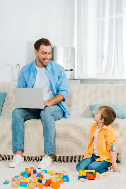 улыбающийся отец сидит на диване и использует ноутбук, в то время как дошкольный сын играет с красочными блоками дома
 - Фото, изображение