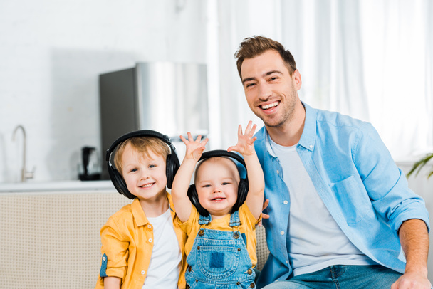 χαμογελαστός ο πατέρας βλέπει κάμερα με γιους αξιολάτρευτο παιδί και preschooler ακουστικά στο σπίτι  - Φωτογραφία, εικόνα