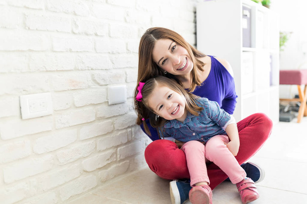 Cheerful mother and daughter wearing casuals while sitting near wall at home - Foto, Bild