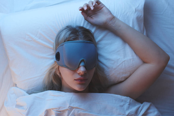 Sleeping girl in a sleep mask on an orthopedic pillow under night lighting, white bedclothes, top view - Foto, imagen