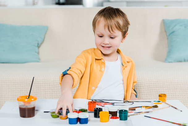 adorable smiling preschooler boy picking up paints during drawing at home - Foto, Imagem