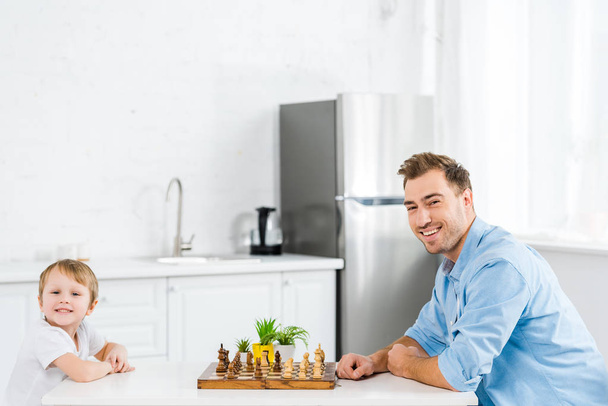 happy father and preschooler son sitting at table and looking at camera while playing chess in kitchen - Фото, изображение