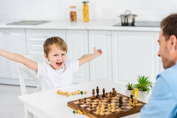 preschooler son cheering with hands in air while playing chess with father at home - Fotó, kép