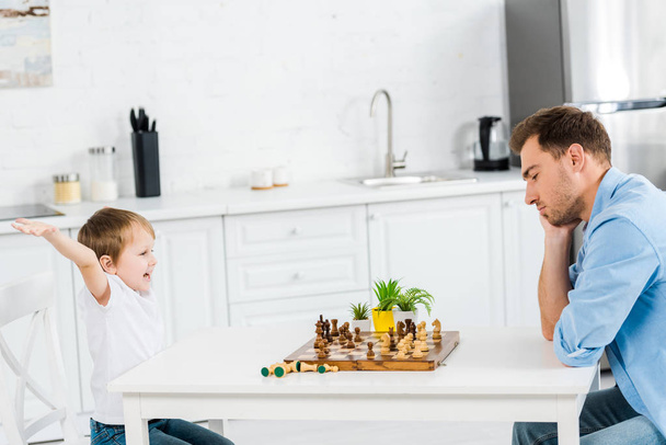 preschooler son cheering with hands in air while playing chess with father at home - Fotografie, Obrázek