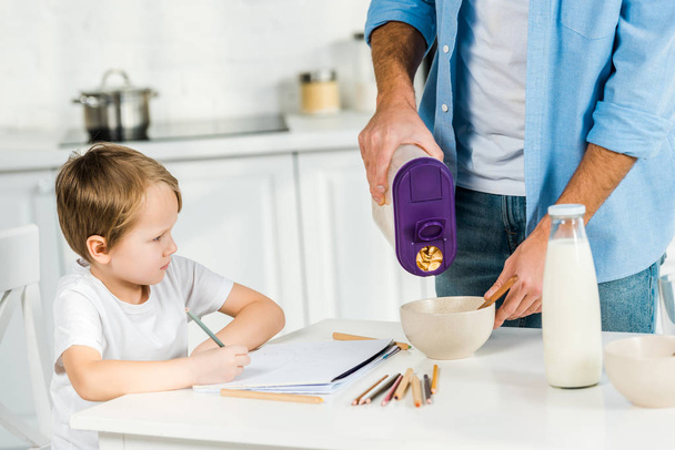 father pouring cereal in bowl while preschooler son drawing during breakfast in morning - Photo, Image