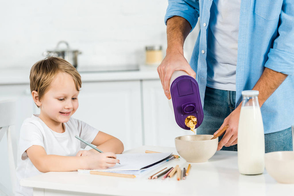 father pouring cereal in bowl while smiling preschooler son drawing during breakfast in morning - Φωτογραφία, εικόνα