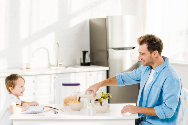 père versant du lait dans un bol tandis que le dessin préscolaire pendant le petit déjeuner dans la cuisine
 - Photo, image