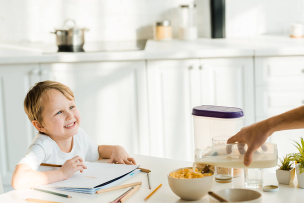 счастливый дошкольник сын рисунок в то время как отец наливать молоко в миску во время завтрака на кухне
 - Фото, изображение