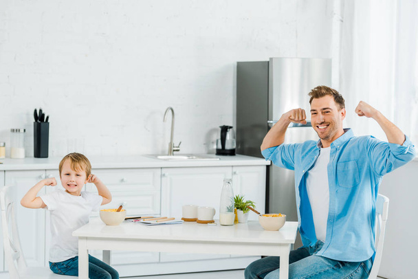 smiling preschooler son and father looking at camera, posing and showing biceps during breakfast in kitchen  - Photo, Image