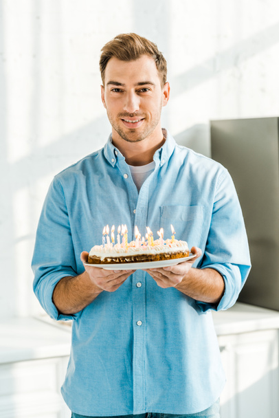 handsome man looking at camera and holding birthday cake with burning candles at home in morning - Foto, immagini