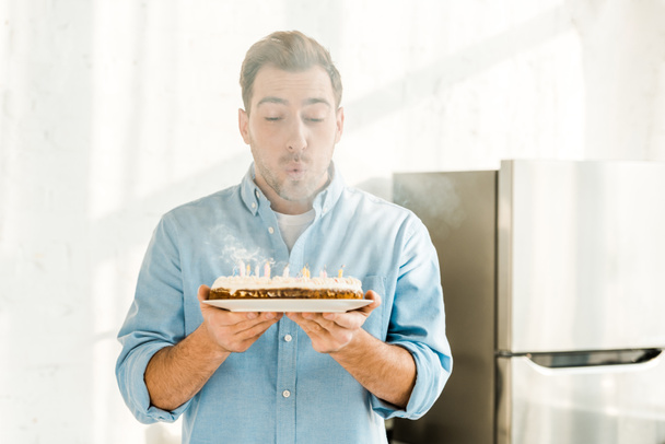 man blowing out candles while holding birthday cake at home in morning  - Foto, immagini