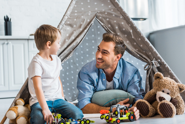 smiling father and preschooler son playing with toy cars and teddy bears under wigwam at home - Valokuva, kuva