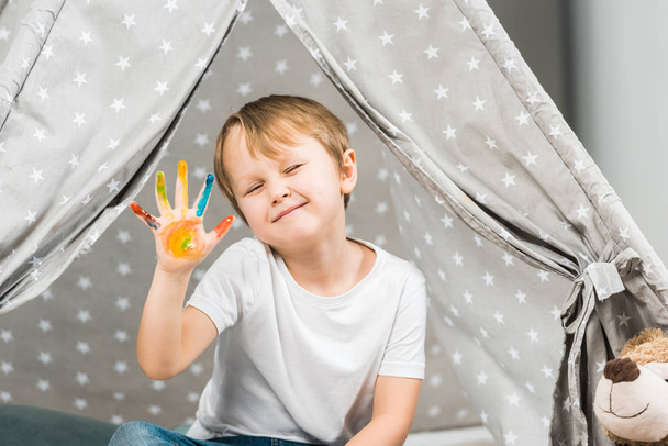 adorable preschooler boy with colorful paint on hand sitting in wigwam at home - Photo, Image