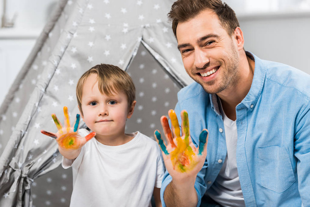 happy father and son looking at camera and showing painted hands at home - 写真・画像