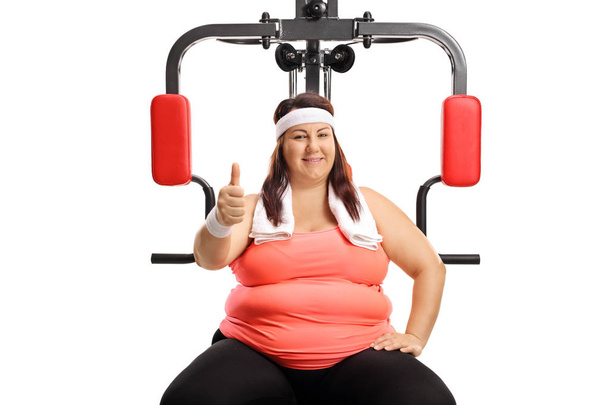 Overweight young woman sitting on a multifunctional gym machine and showing thumbs up isolated on white background - Foto, Imagem