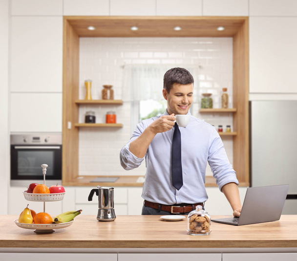 Przystojny młody mężczyzna, picia kawy i działa na laptopie w swojej kuchni - Zdjęcie, obraz
