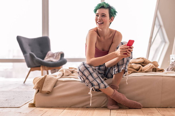 Green-haired woman sitting on bed and using red smartphone - Photo, Image