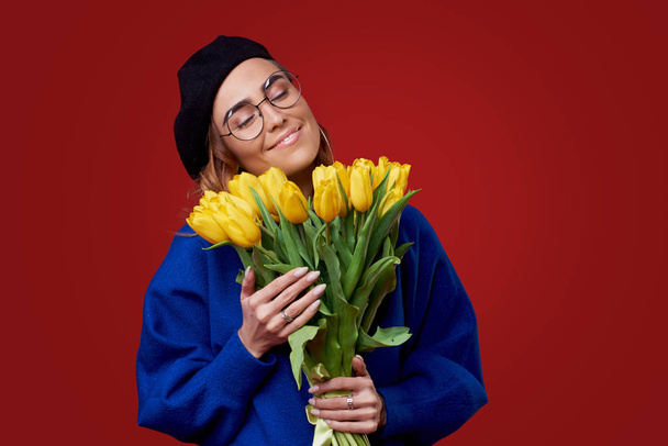 Cheerful young lady in blue clothes, black beret and round glasses being excited to get bouquet of yellow tulips on women's day or birthday isolated over red background. French style girl  - Photo, Image