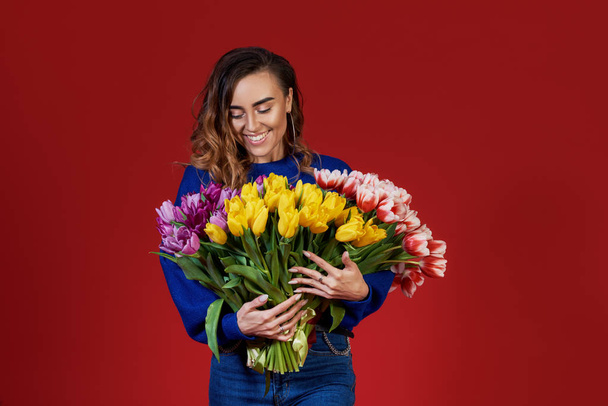 Cheerful young lady in blue clothes, holding and smelling bouquet of colorful tulips on women's day or birthday isolated over red background.  - Zdjęcie, obraz
