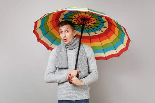 Σοκαρισμένος νεαρός άνδρας σε γκρι πουλόβερ, κασκόλ κρατώντας πολύχρωμες ομπρέλες - Φωτογραφία, εικόνα