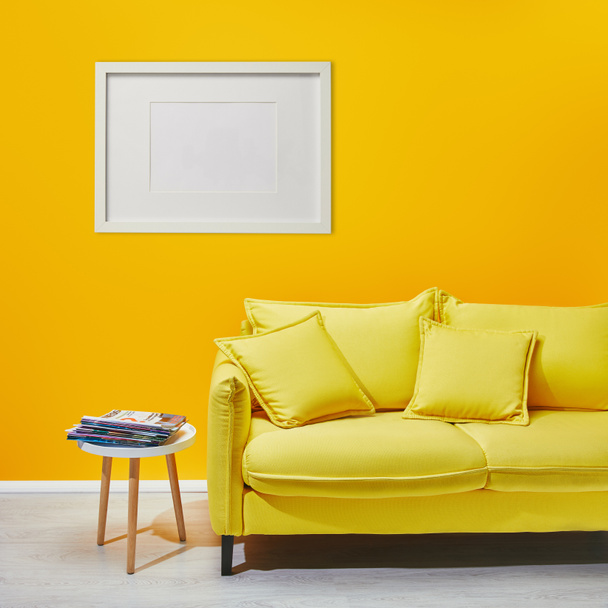 coffee table standing near modern yellow sofa near white frame hanging on wall - Foto, afbeelding