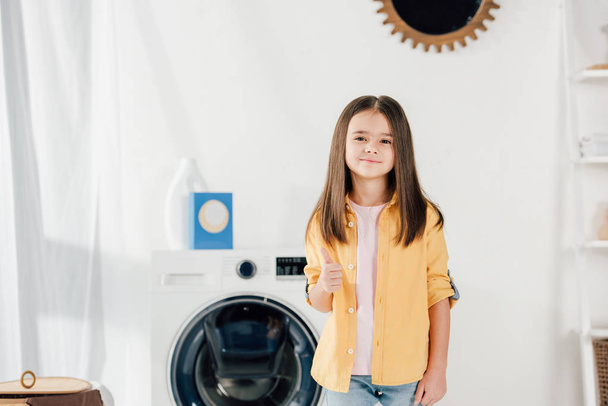 Kind in gelbem Hemd und Jeans steht und zeigt Daumen hoch in Waschküche - Foto, Bild