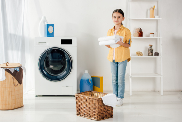 child in yellow shirt near washer and ladder holding towels in laundry room - Foto, Imagen