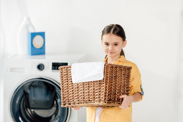 παιδί σε κίτρινο πουκάμισο κρατώντας καλάθι με πετσέτα στο δωμάτιο πλυντηρίων - Φωτογραφία, εικόνα