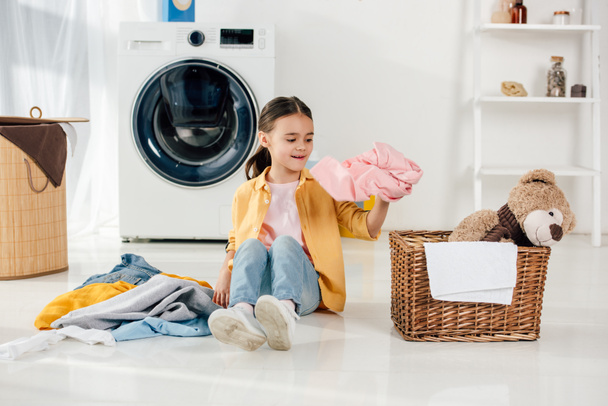niño en camisa amarilla y jeans sentado y poniendo ropa a la cesta en la sala de lavandería
 - Foto, imagen