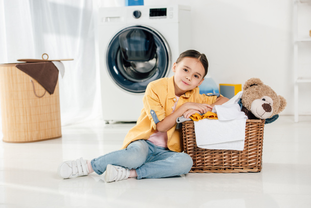 niño en camisa amarilla y jeans sentado cerca de la cesta con juguete de oso en la sala de lavandería
 - Foto, Imagen