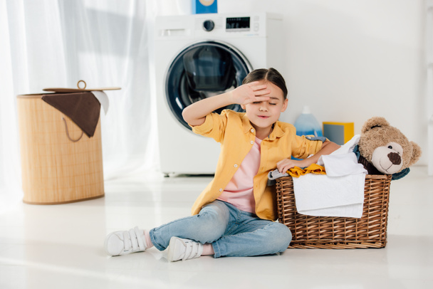 Müdes Kind in gelbem Hemd und Jeans sitzt neben Korb mit Bärenspielzeug in Waschküche - Foto, Bild