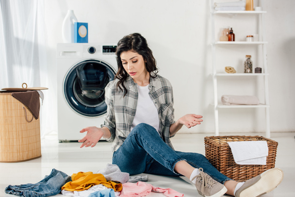 Verwirrte Frau sitzt auf dem Boden neben verstreuten Kleidungsstücken und Körben in Waschküche - Foto, Bild