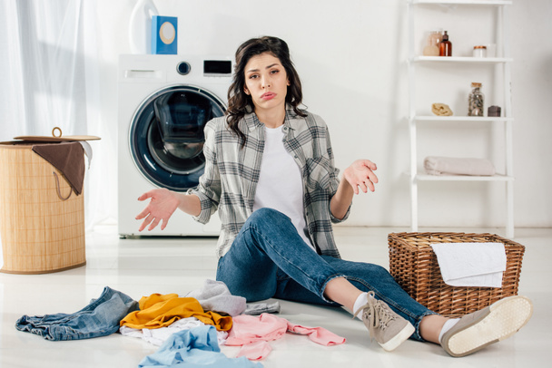 Müde Frau sitzt auf dem Boden neben verstreuten Kleidungsstücken und Körben in Waschküche - Foto, Bild