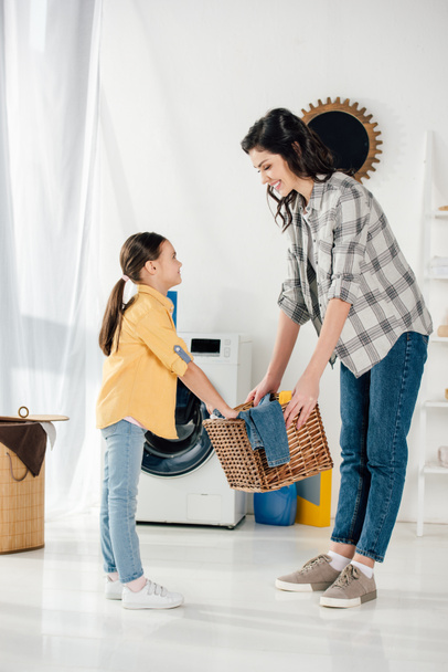 daughter in yellow shirt and mother in grey shirt holding basket and smiling in laundry room - Foto, afbeelding