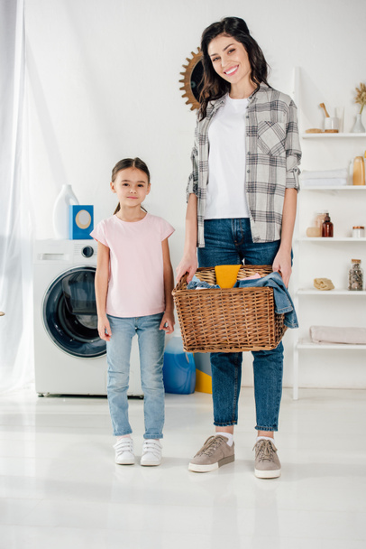 dochter in roze t-shirt staande in de buurt van lachende moeder in grijs shirt met mand en wasruimte - Foto, afbeelding