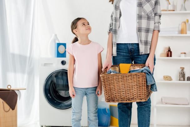 Tochter im rosa T-Shirt schaut Mutter im grauen Hemd mit Korb in Waschküche an - Foto, Bild