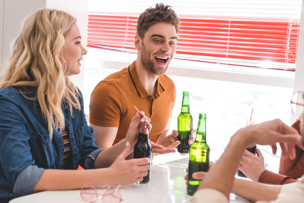 amigos sonrientes sosteniendo botellas de vidrio con bebida y hablando en la cafetería
  - Foto, Imagen