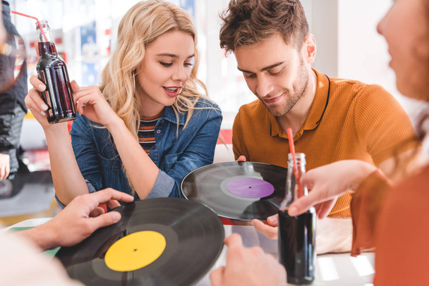 selective focus of handsome and beautiful friends talking, holding vinyl and glass bottles with drink  - Φωτογραφία, εικόνα