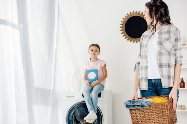 selective focus of daughter sitting on washer with washing powder wile mother holding basket in laundry room - Foto, afbeelding