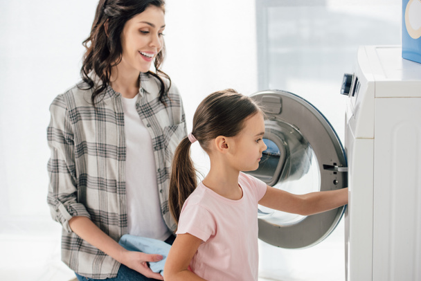 daughter and mother sitting near opened washer in laundry room - Φωτογραφία, εικόνα