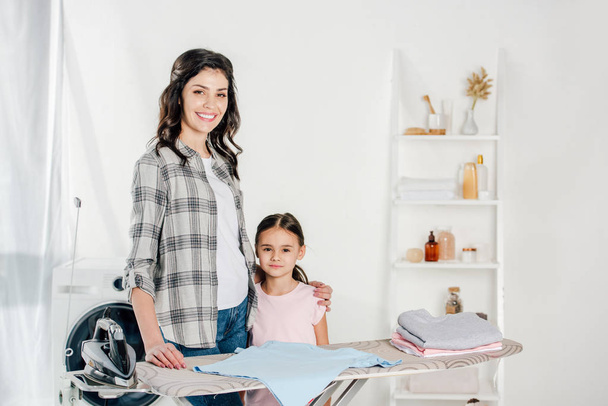 mother and daughter standing near ironing board in laundry room - Photo, Image