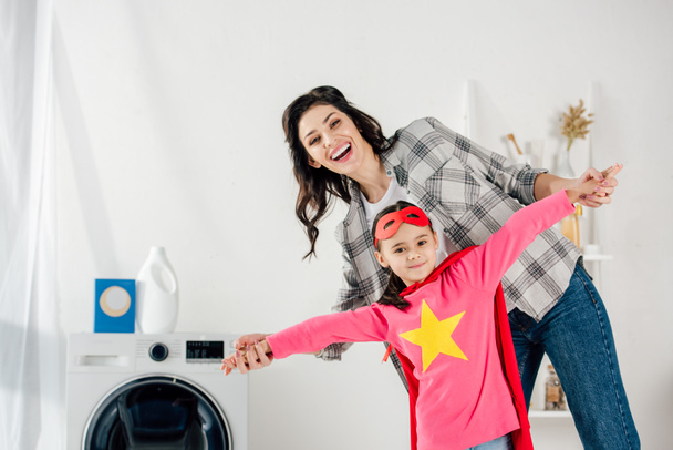 moeder in grijs shirt en dochter in rode zelfgemaakte pak met sterrenbeeld plezier in wasruimte - Foto, afbeelding