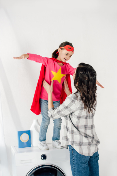 mother in grey shirt touching daughter in red homemade suit with star sign on washer in laundry room - Φωτογραφία, εικόνα