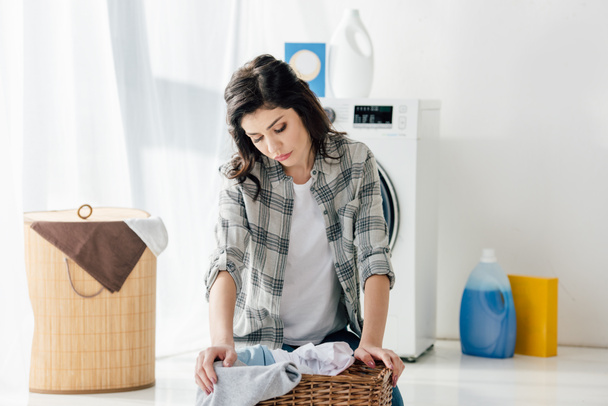 Aufgebrachte Frau im grauen Hemd sitzt neben Korb in Waschküche - Foto, Bild