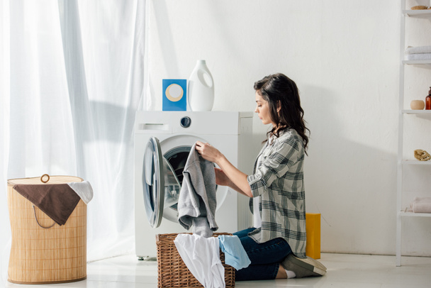 Frau in grauem Hemd und Jeans legt Kleidung in Korb neben Waschmaschine in Waschküche - Foto, Bild