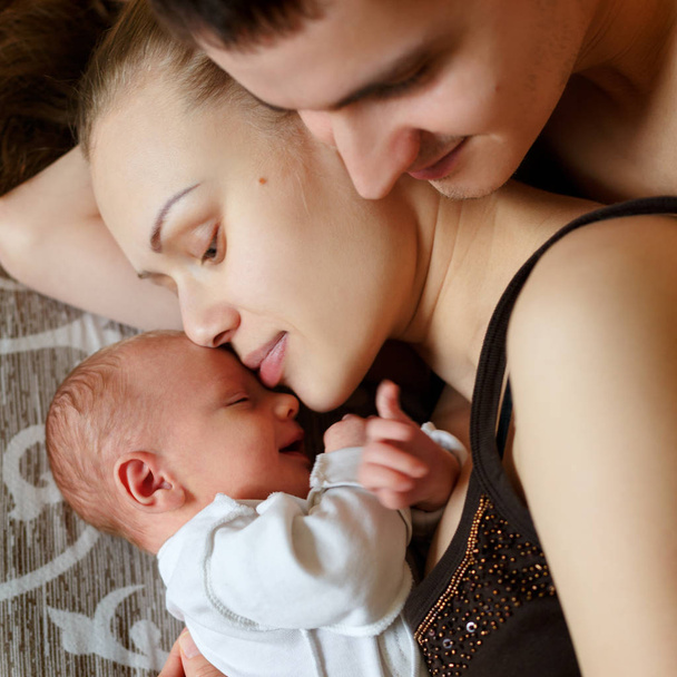 Woman and man lie on bed together with newborns. Mom, dad and baby. Boy clung to his mother. Portrait of young smiling family with newborn. Happy family life. Man was born. - Foto, immagini