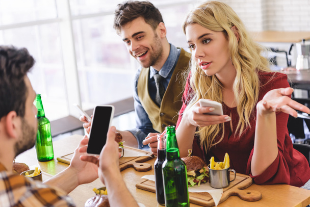 Selektivní fokus krásných a pohledný přátel mluví a používají smartphony v kavárně  - Fotografie, Obrázek