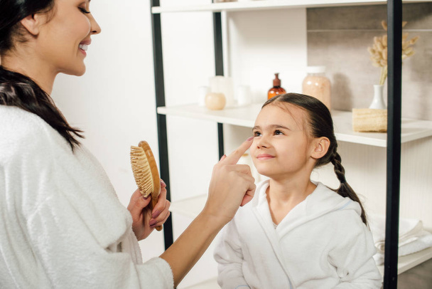 mother holding hairbrush and touching daughter nose in bathroom - Фото, изображение