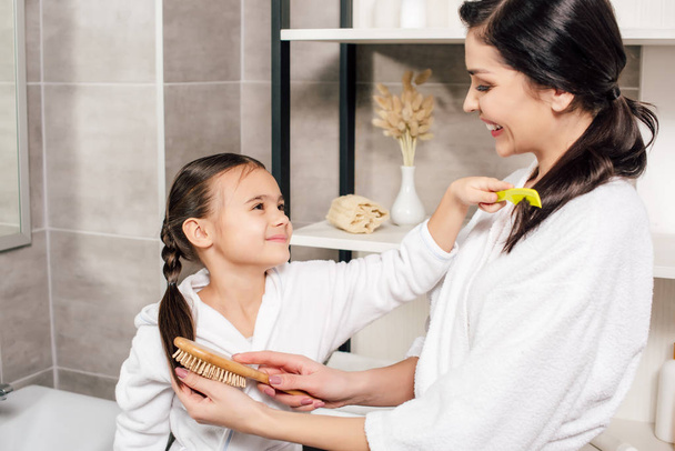 mother and daughter in white bathrobes combing each other in bathroom - Photo, Image