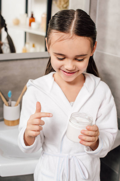 child in white bathrobe holding container with cosmetic cream in bathroom - Photo, image